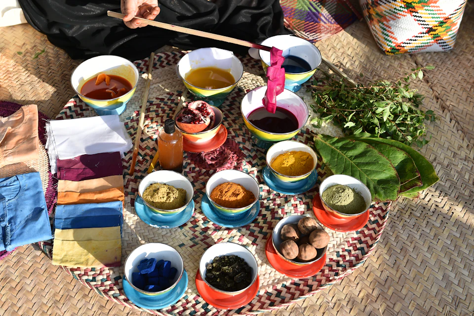 A collection of colorful natural dye ingredients and pigments in bowls, a person's hand holding a stick with dyed fabric.