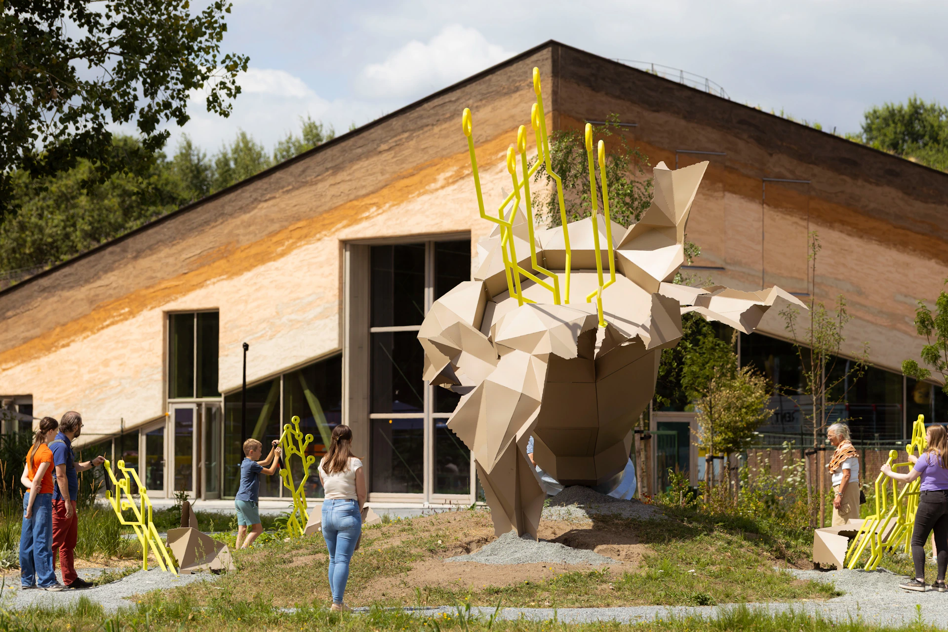 A geometric sculpture with yellow rods in front of a modern building, with people observing the artwork outdoors.