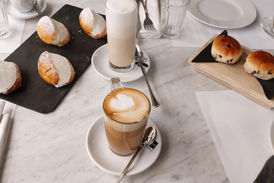 Two cups of coffee on a marble table, surrounded by pastries, utensils, a glass, and plates.