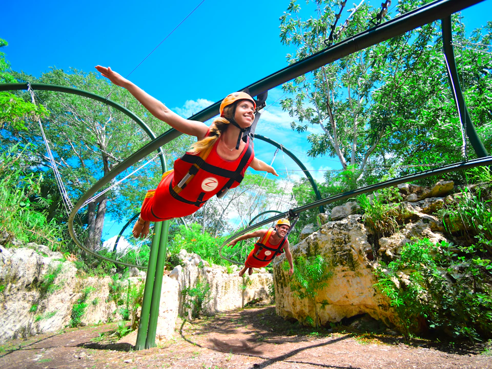People in bright red harnesses glide through a canopy on a zip line track in a lush, rocky environment on a sunny day.