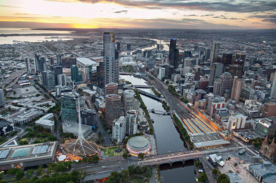 Aerial view of a cityscape featuring skyscrapers, a winding river with bridges, and a sunset sky in the background.