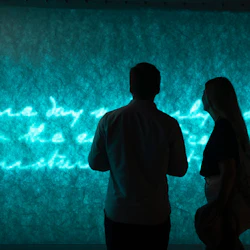 Två personer i silhuett står framför en glödande, turkos vägg med kursiv text.