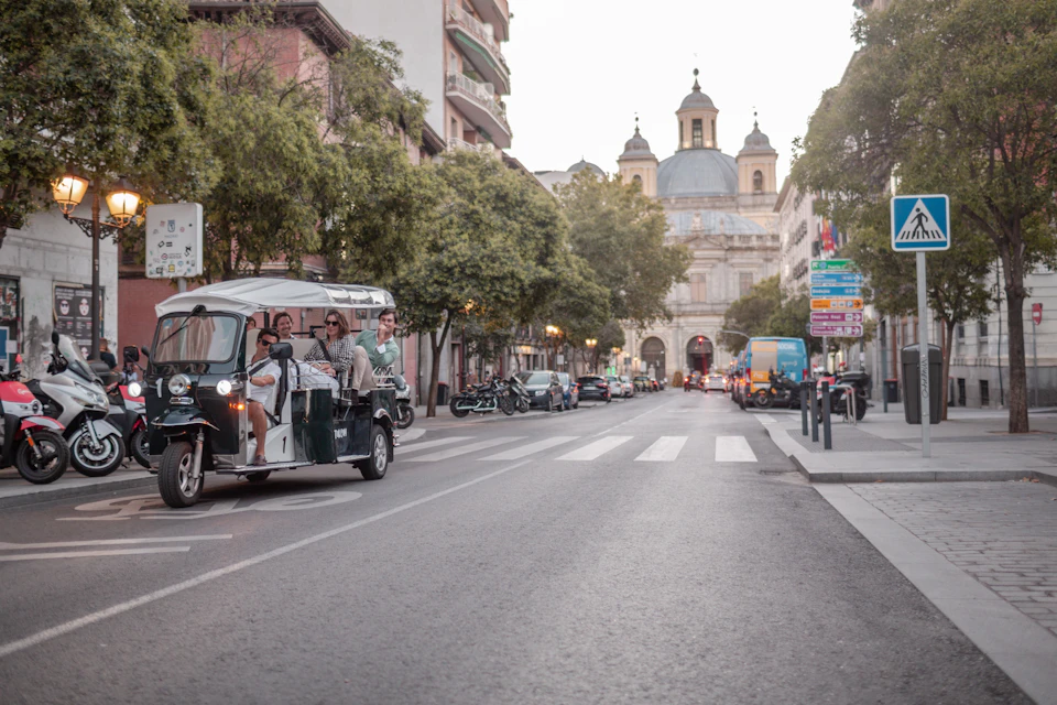 A street scene with people riding in a tuk-tuk, cars parked along the sides, trees, buildings, and a large church in the background.