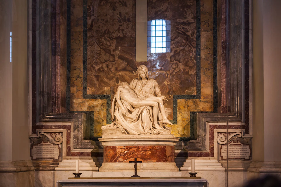 A marble statue depicting a seated figure cradling a reclining figure, set in a niche with ornate marble walls and a small cross at the front.