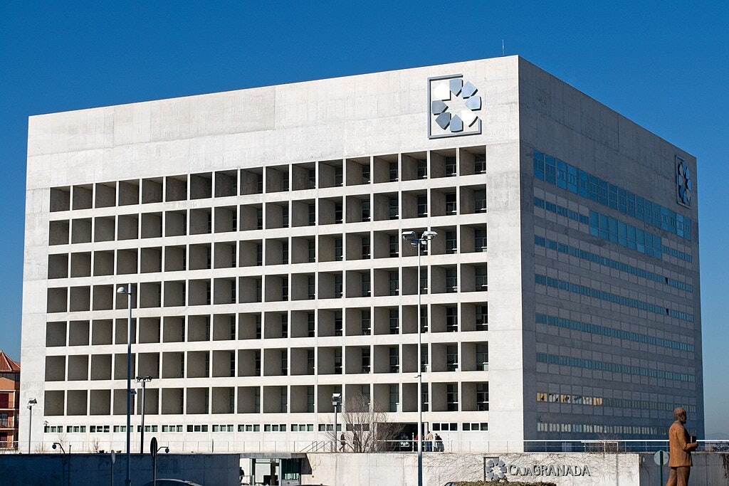 Un grand immeuble de bureaux moderne avec une façade en forme de grille et un logo dans les coins supérieurs, sous un ciel bleu clair.