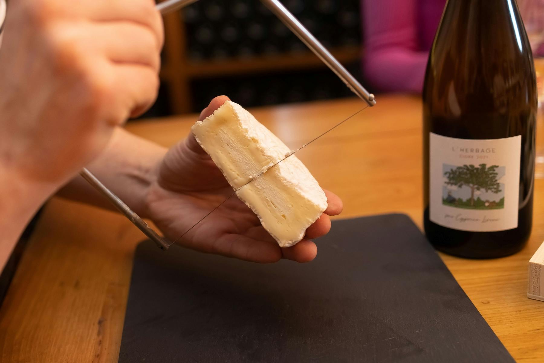 Рука, нарезающая кусок сыра проволочным сырорезом над черной разделочной доской, с бутылкой на заднем плане.