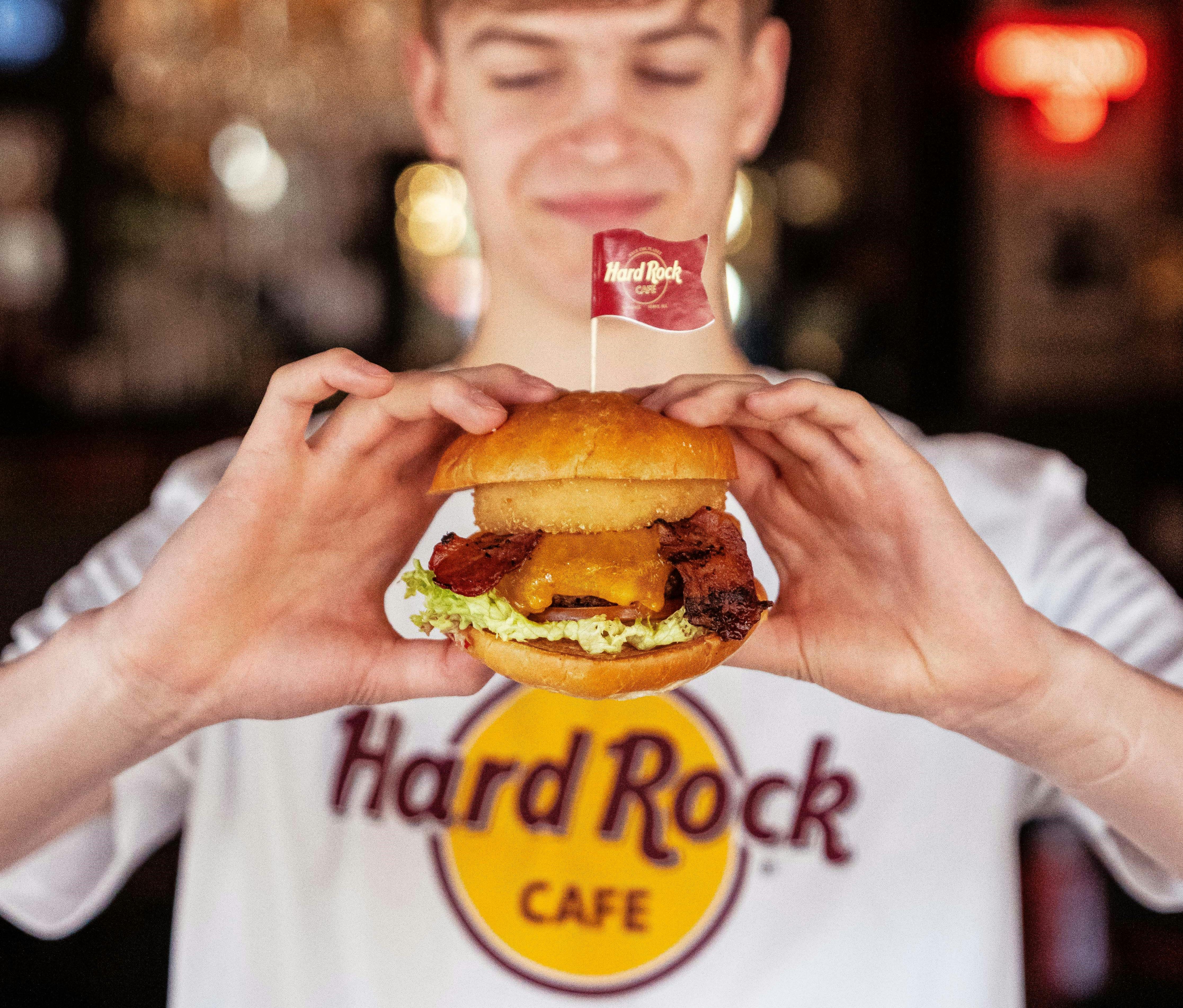 Personne portant une chemise du Hard Rock Cafe et tenant un hamburger avec de la laitue, du bacon et du fromage, surmonté d'un petit drapeau Hard Rock.