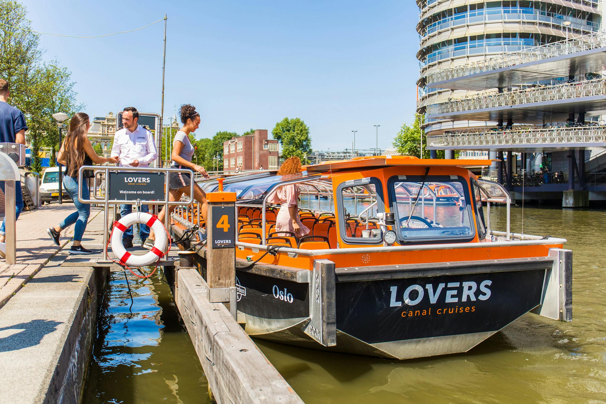 Mensen stappen aan boord van een oranje rondvaartboot aan een steiger met het opschrift "LOVERS" onder een strakblauwe lucht. Bomen en gebouwen op de achtergrond.