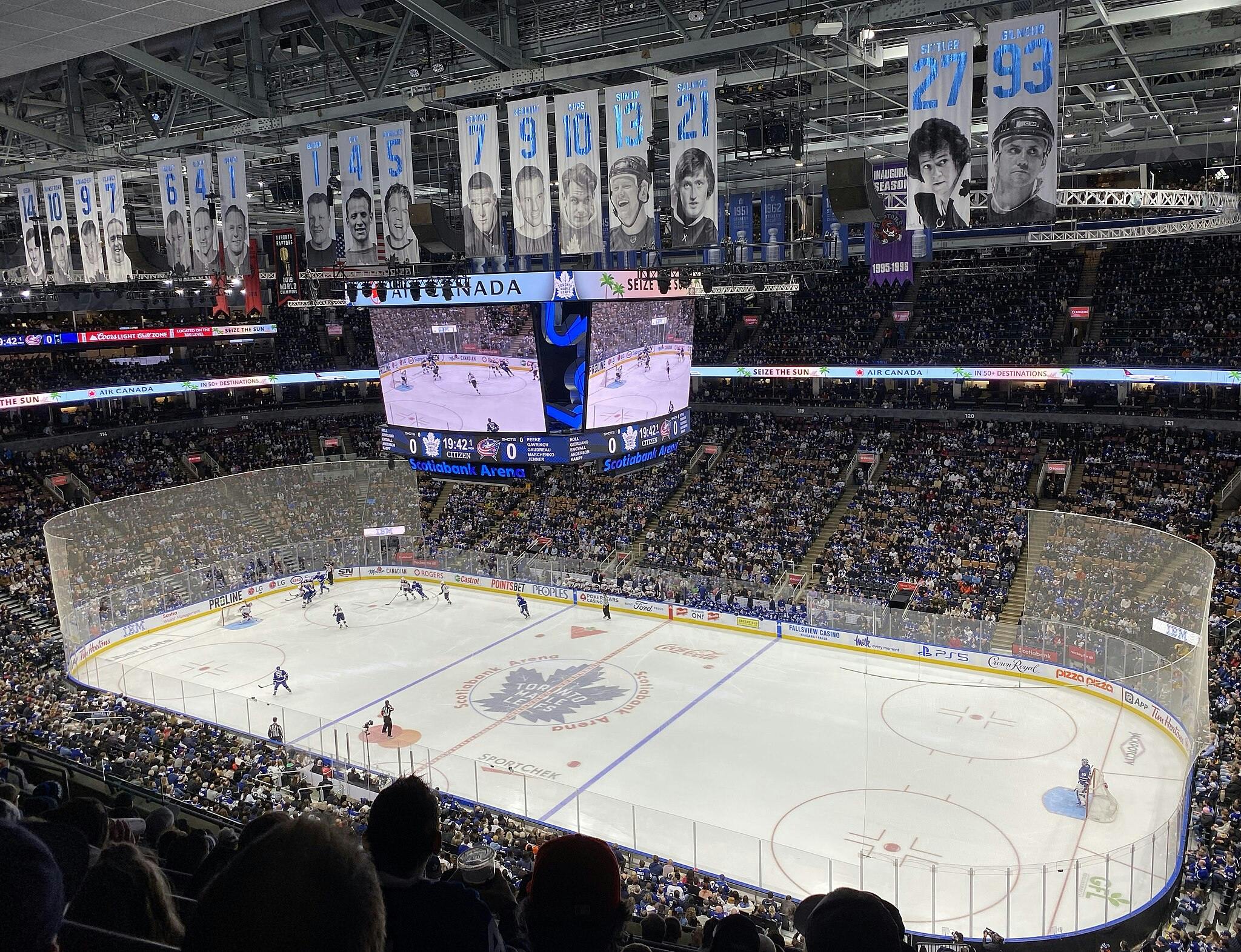 Naplněná krytá aréna s fanoušky, kteří sledují intenzivní hokejový zápas na kluzišti pod bannery vysloužilých hráčů.
