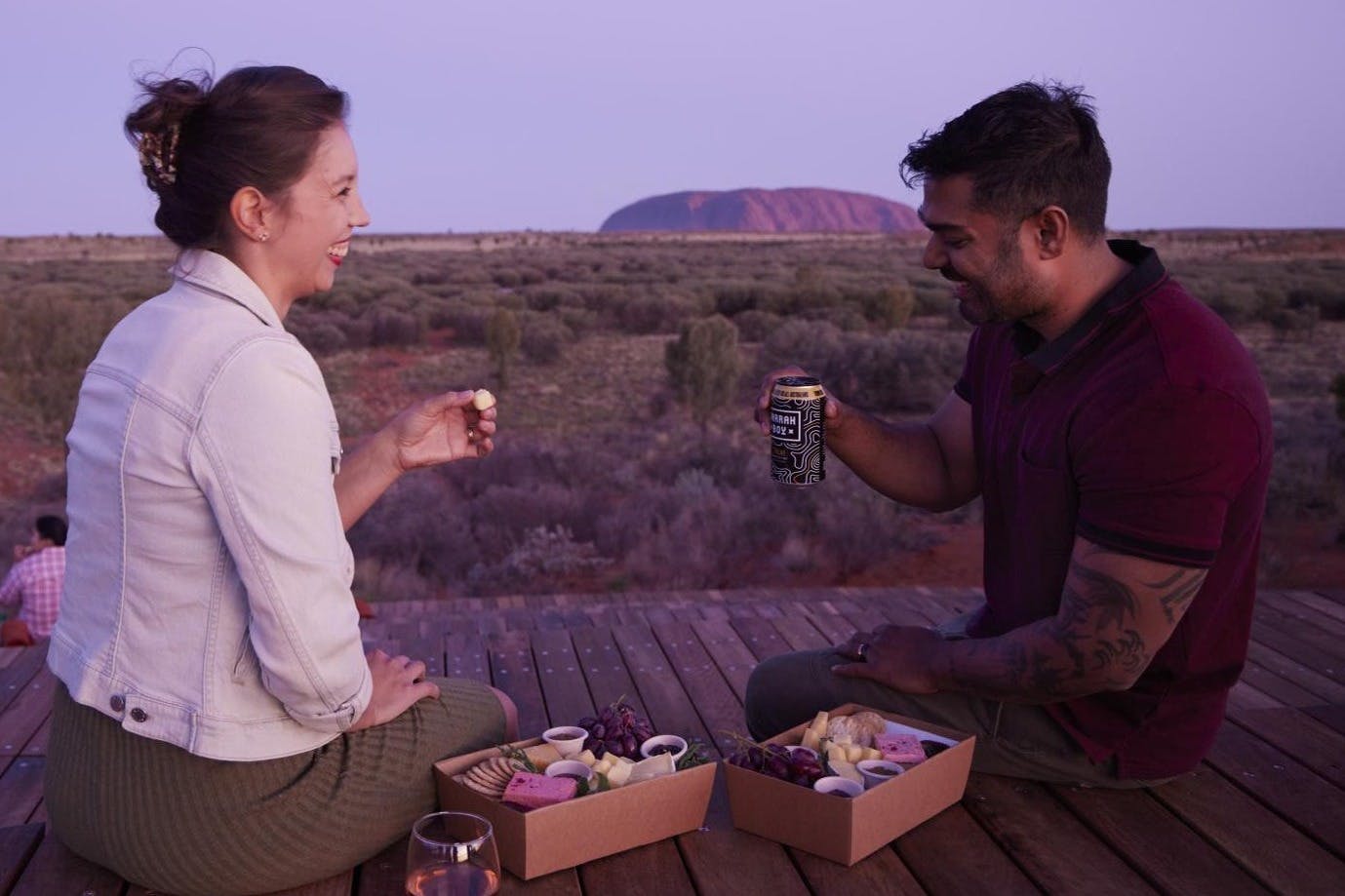 Woman seated on a wooden deck, enjoying a picnic with snacks and drinks at sunset, with a desert landscape and large rock formation in the background.