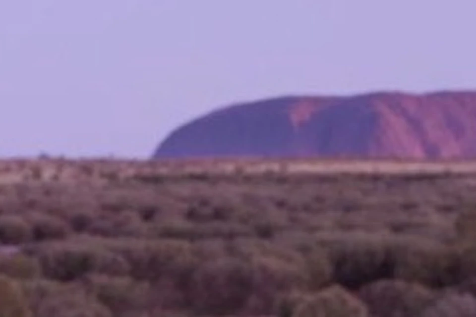 Woman seated on a wooden deck, enjoying a picnic with snacks and drinks at sunset, with a desert landscape and large rock formation in the background.
