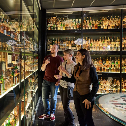 Tre personer undersöker hyllor fyllda med många flaskor whisky i ett väl upplyst utställningsrum.