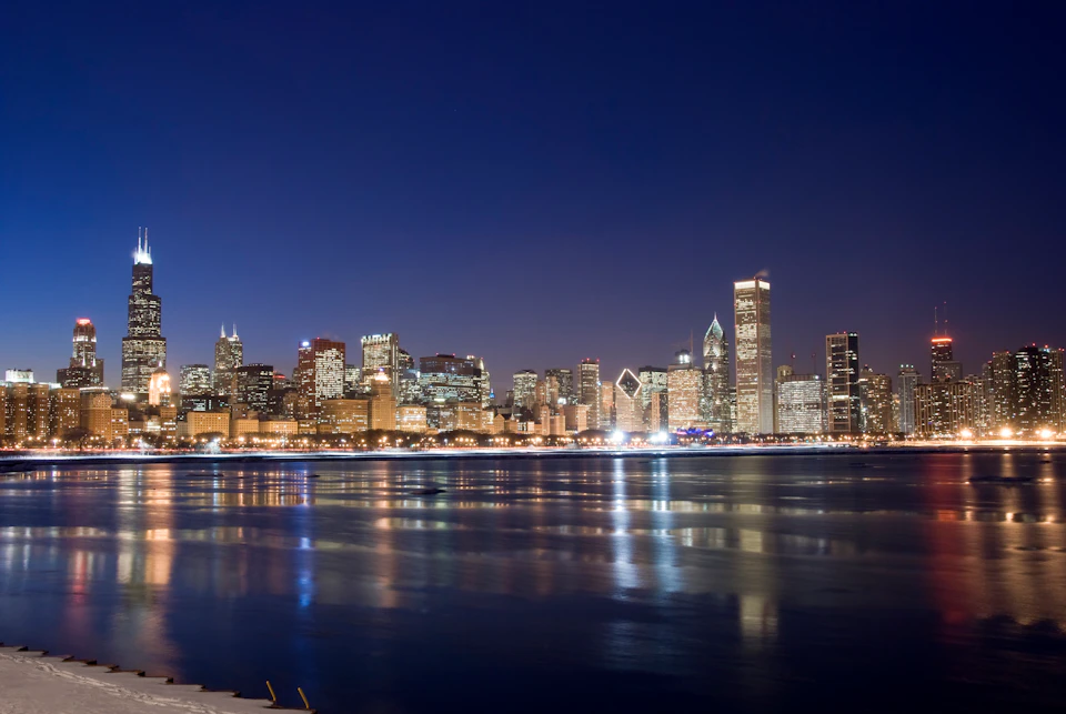 City skyline at night with illuminated buildings reflecting on calm water.
