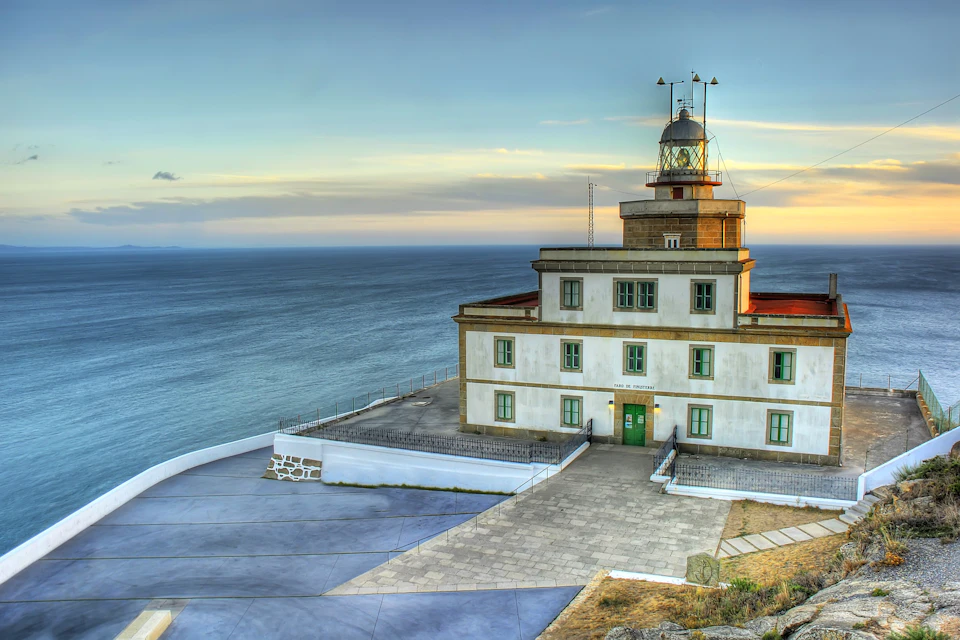 A coastal lighthouse with a rectangular building, situated on a lookout, overlooking a calm ocean under a clear, evening sky.