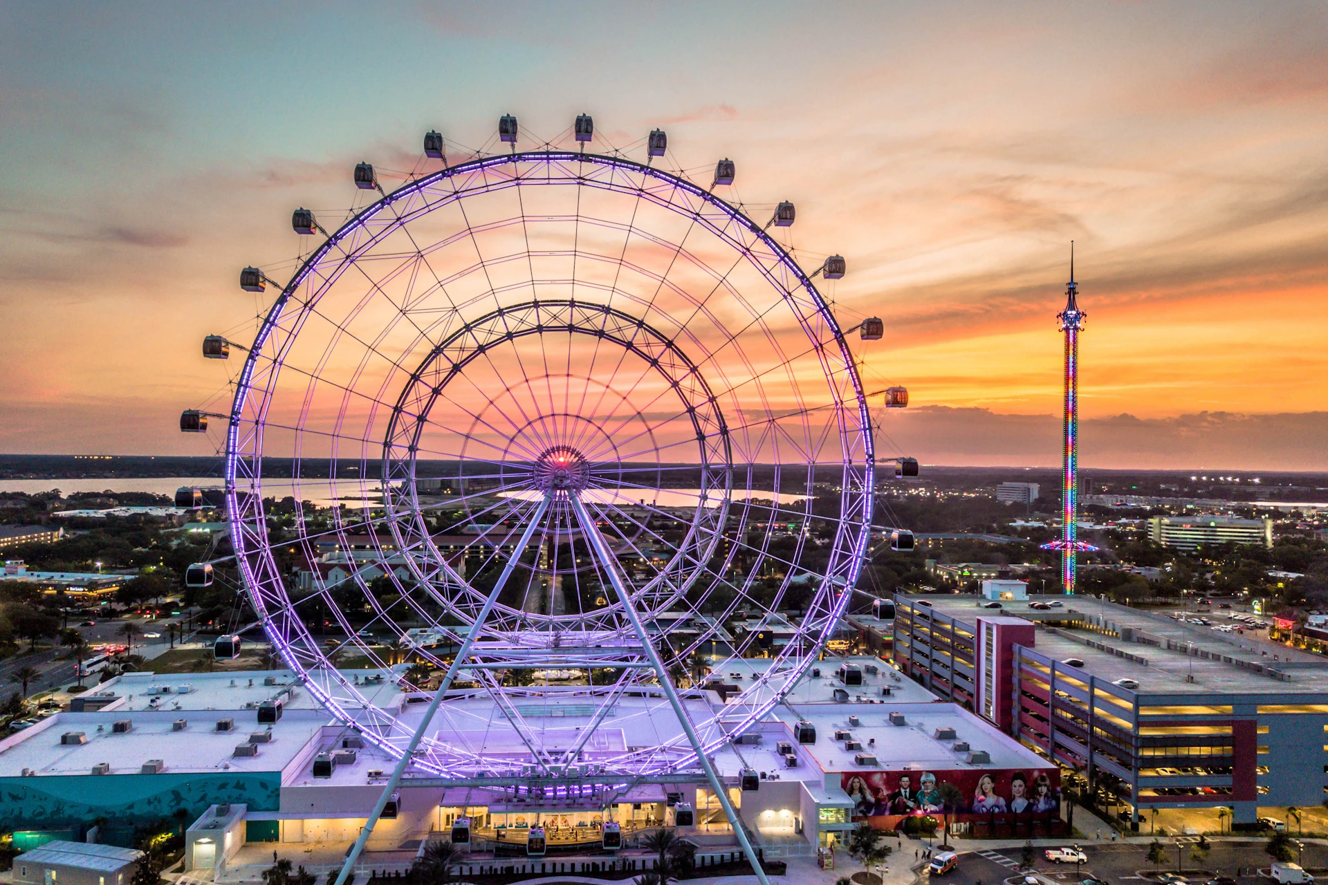 A large ferris wheel lit with purple lights at sunset, with a cityscape and a tall illuminated tower in the background.