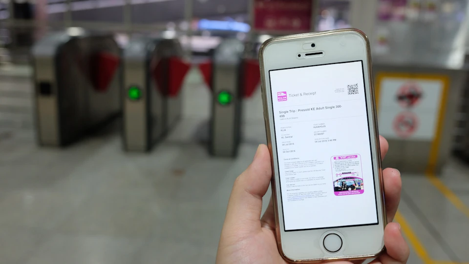 A person holding a smartphone displaying a digital ticket, with electronic turnstiles visible in the background.