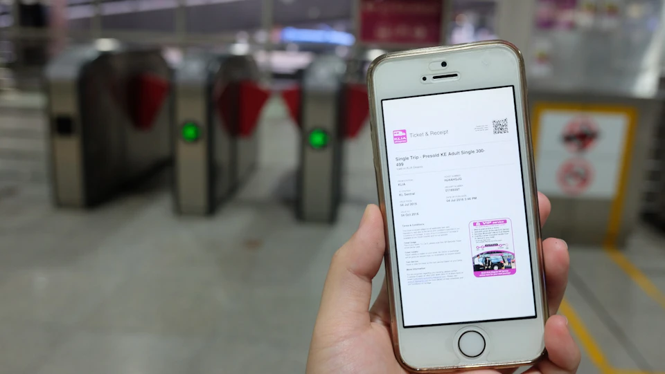 A person holding a smartphone displaying a digital ticket, with electronic turnstiles visible in the background.