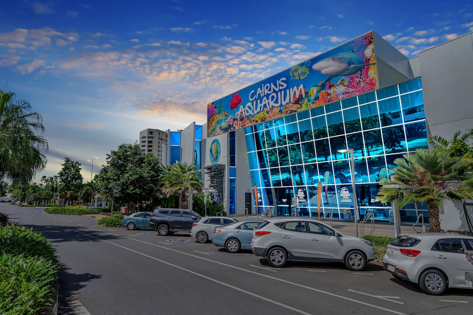 A modern building with large glass windows and a colorful sign reading 'Cairns Aquarium,' with parked cars in front.