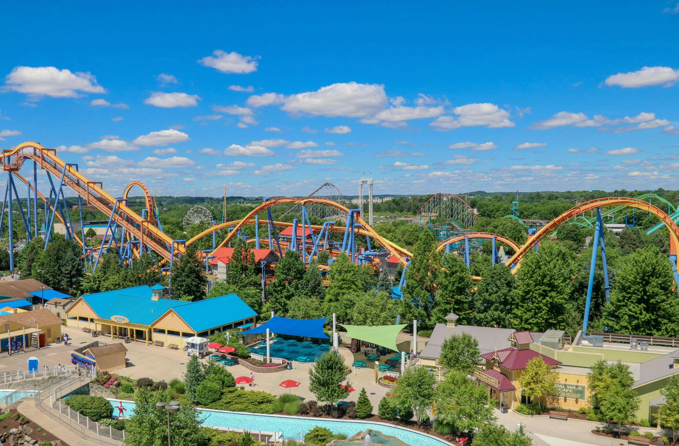 Um parque temático com montanhas-russas coloridas, árvores verdes, edifícios, áreas de estar e um céu azul brilhante com nuvens dispersas.