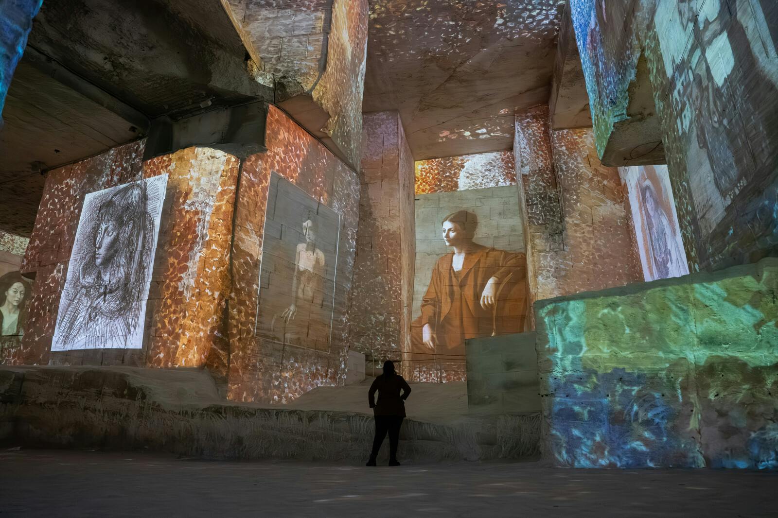 A person observes illuminated artwork projections on the walls of a large space with rock-like surfaces and vibrant colors.