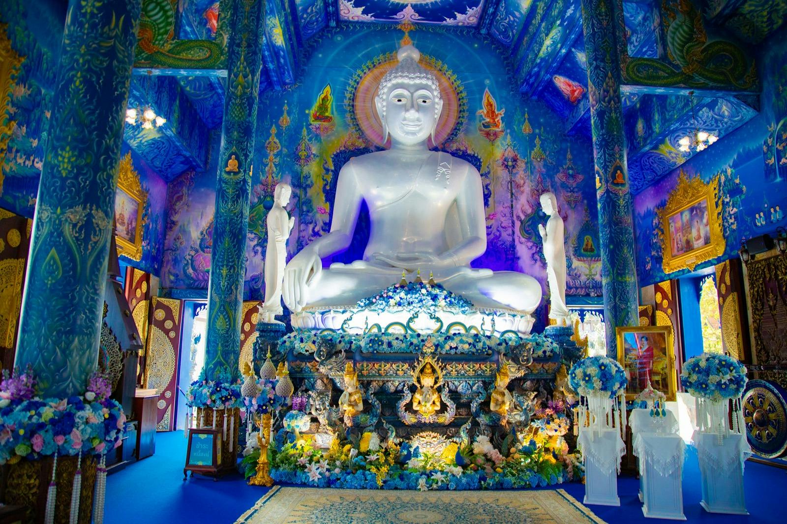 Eine große weiße Buddha-Statue in einem verschnörkelten, leuchtend blauen Tempelinneren mit aufwändigen Dekorationen und Blumenarrangements.