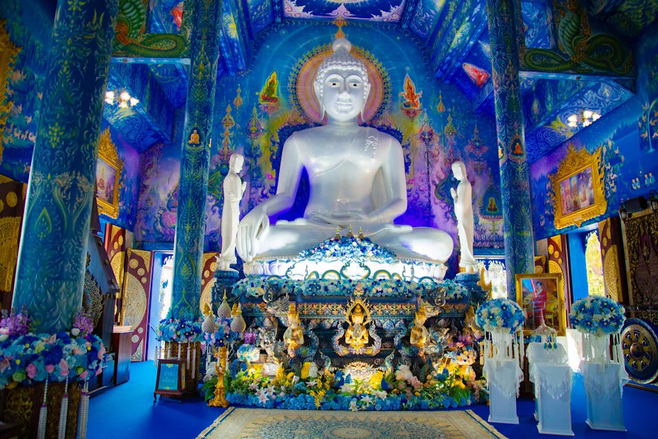 A large white Buddha statue in an ornate, vibrant blue temple interior with intricate decorations and floral arrangements.