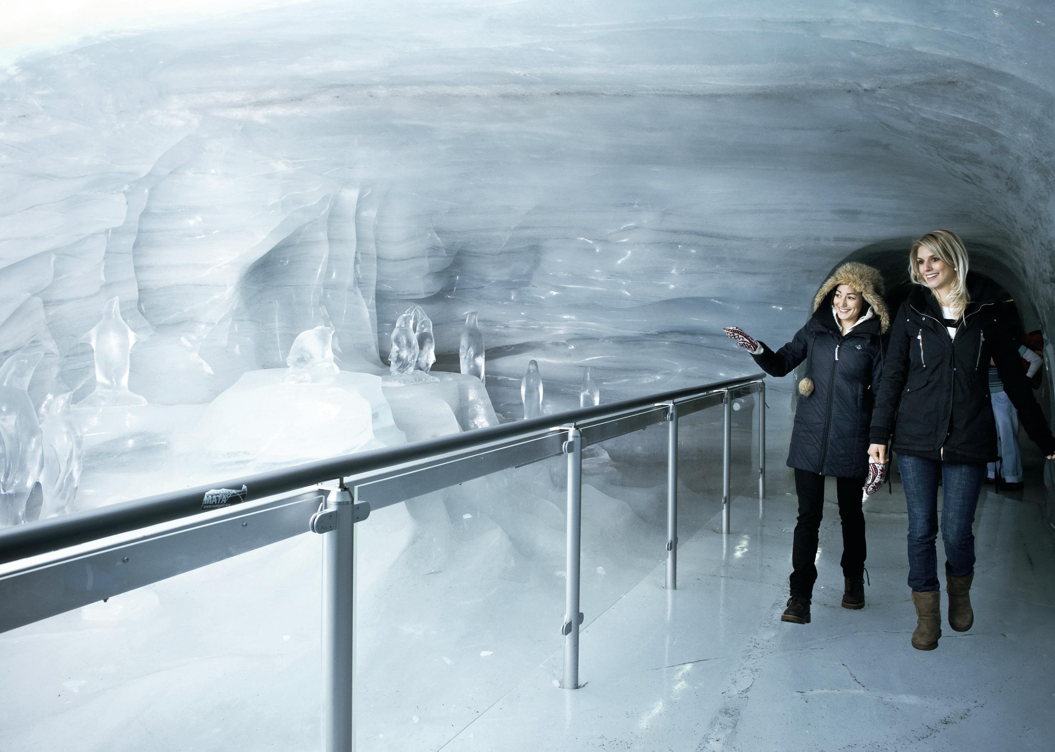 冬のコートを着た2人の女性が氷のトンネルを歩いていきます。左側には手すりがあり、笑顔で話をしています。