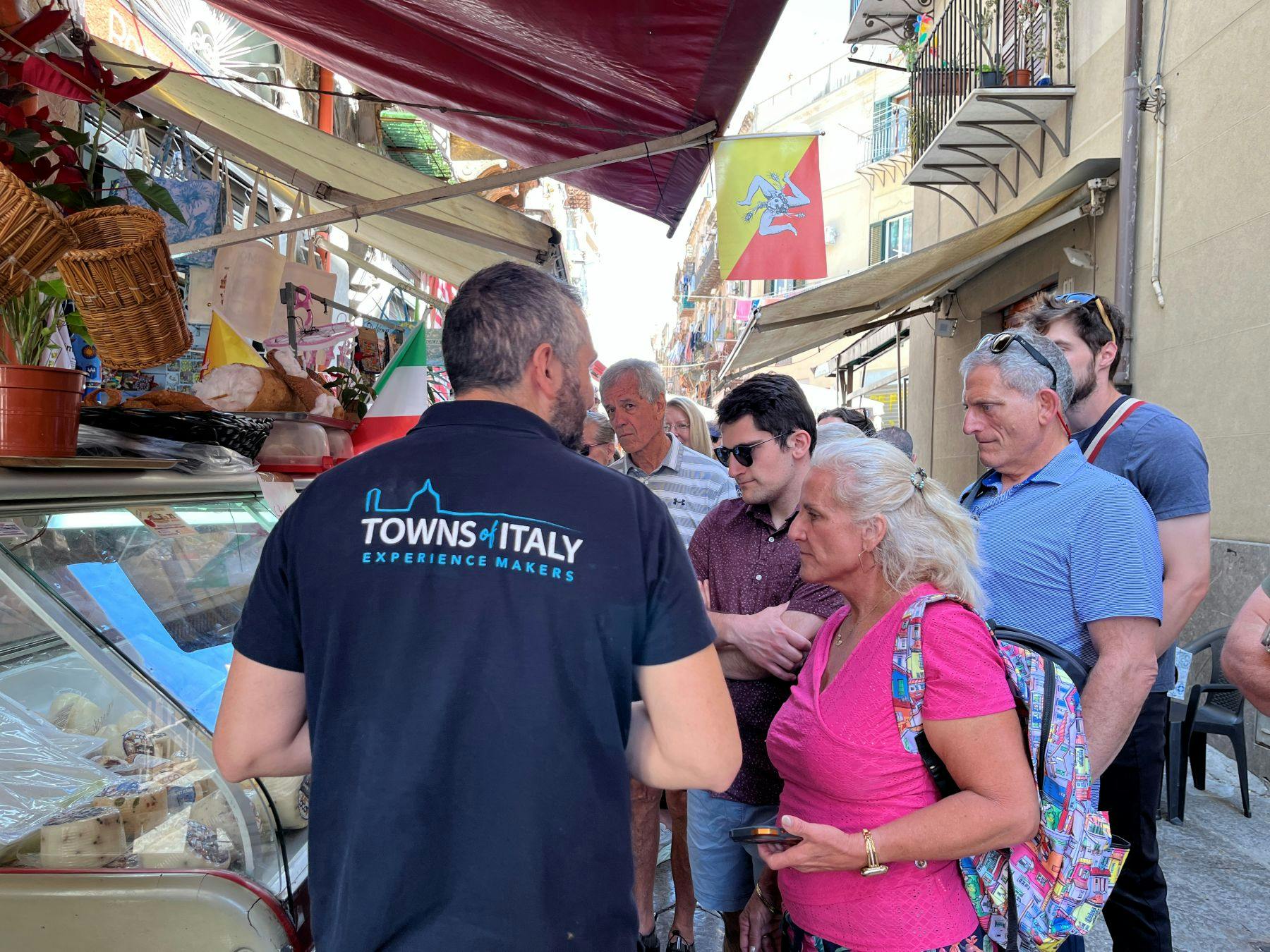 A tour guide interacts with a group of people on a street, with flags and buildings visible in the background.