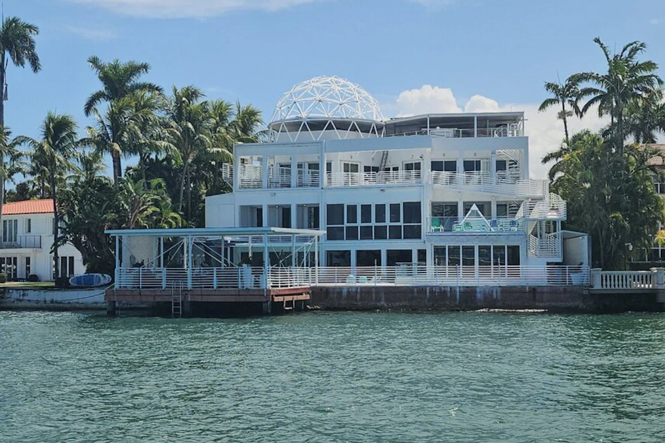 Modern white waterfront building with multiple balconies and a geodesic dome on top, surrounded by palm trees.