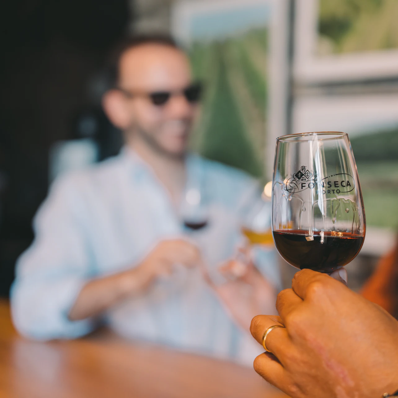 A person holding a glass of red wine in focus, with a blurred background showing a person in sunglasses at a counter.