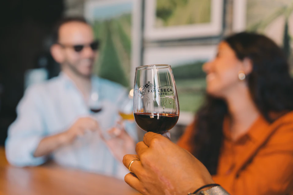 A person holding a glass of red wine in focus, with a blurred background showing a person in sunglasses at a counter.