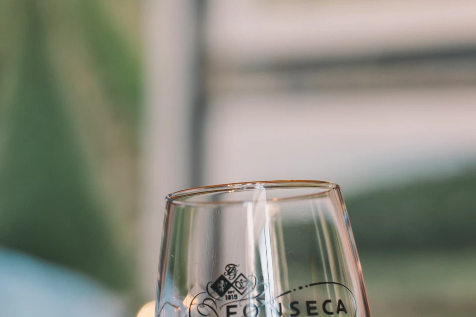A person holding a glass of red wine in focus, with a blurred background showing a person in sunglasses at a counter.