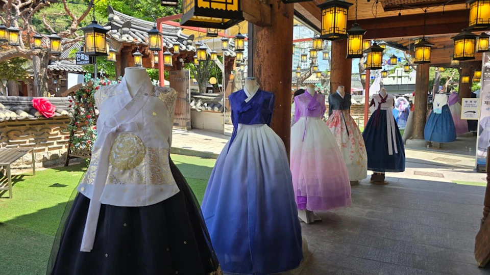 Mannequins displaying traditional Korean hanbok dresses in a market setting with lanterns and wooden columns.