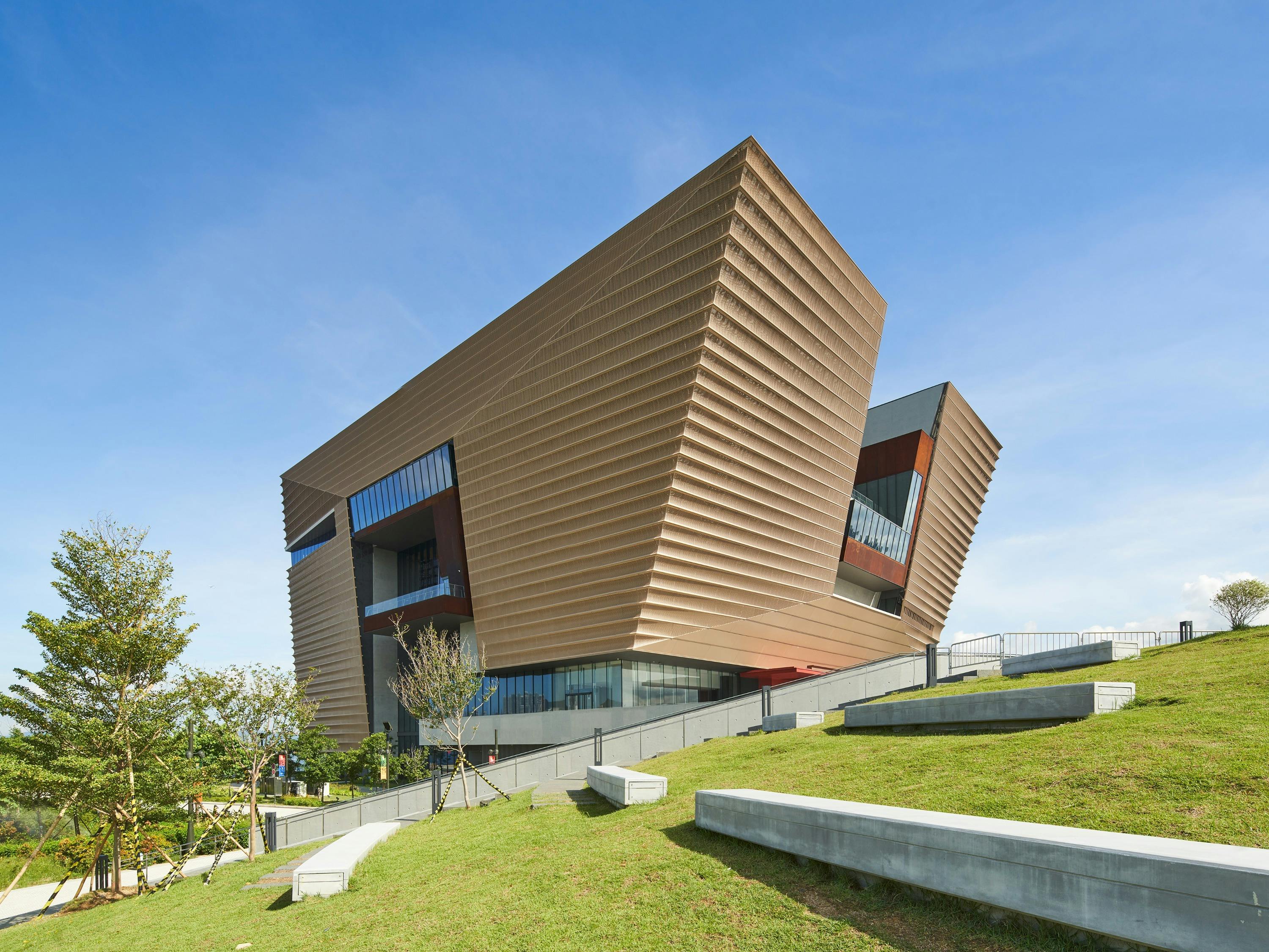 A modern architectural building with angular, textured walls, set on a grassy slope under a clear blue sky.