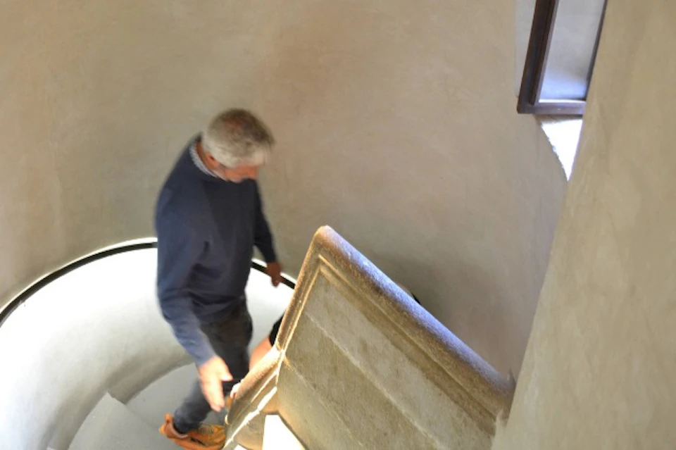 A man descending a spiral staircase inside a building with beige walls and a narrow window visible on the right.