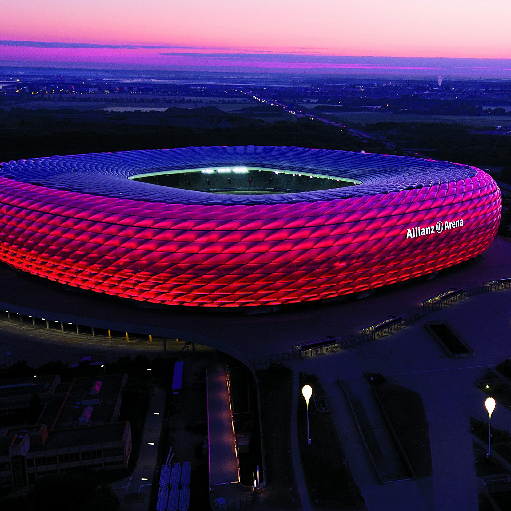 Stadion s červeně osvětleným exteriérem za soumraku, označený jako „Allianz Arena“, obklopený tmavou krajinou pod růžovo-fialovou oblohou.