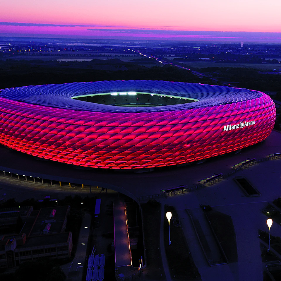 Stadium with red illuminated exterior at dusk, labeled "Allianz Arena," surrounded by dark landscape under a pinkish-purple sky.