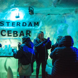 Människor i vinterkappor njuter av drinkar i en svagt upplyst isbar med "Amsterdam Icebar" på en frostig vägg.