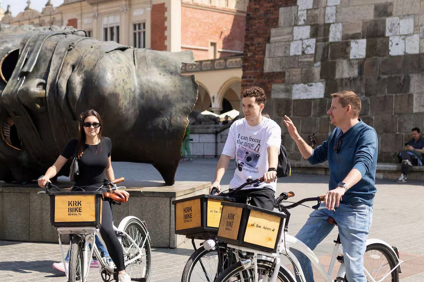 歴史的建造物と大きな彫刻が背景にある公共広場で、自転車の隣に立って会話をしている2人の人物。