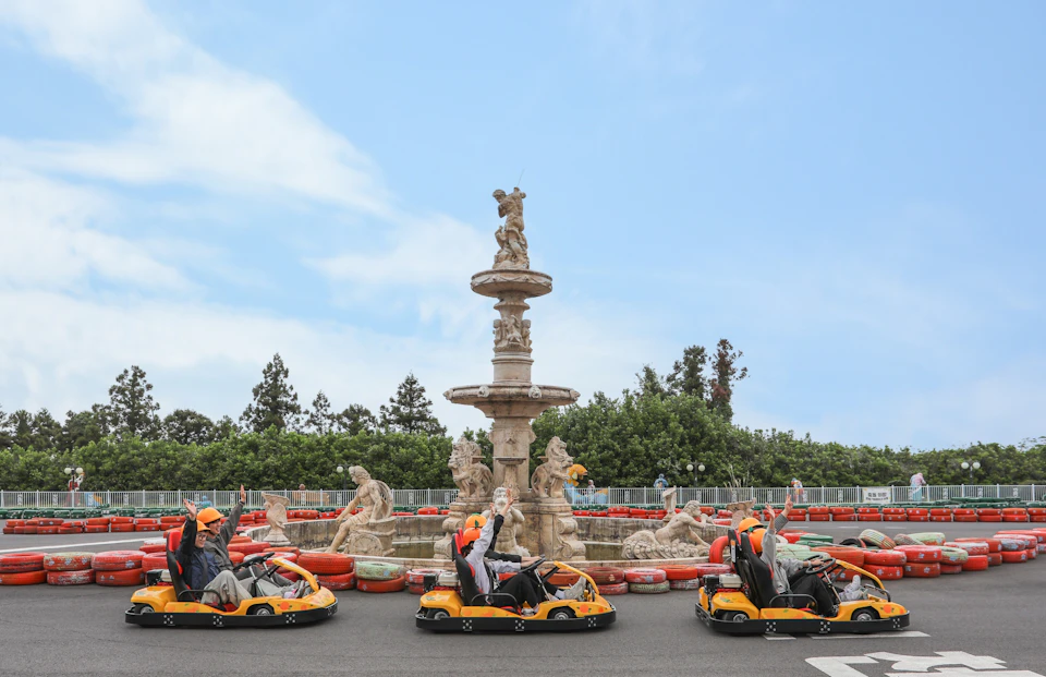 People driving yellow go-karts in front of a decorative stone fountain with sculptures, under a blue sky.