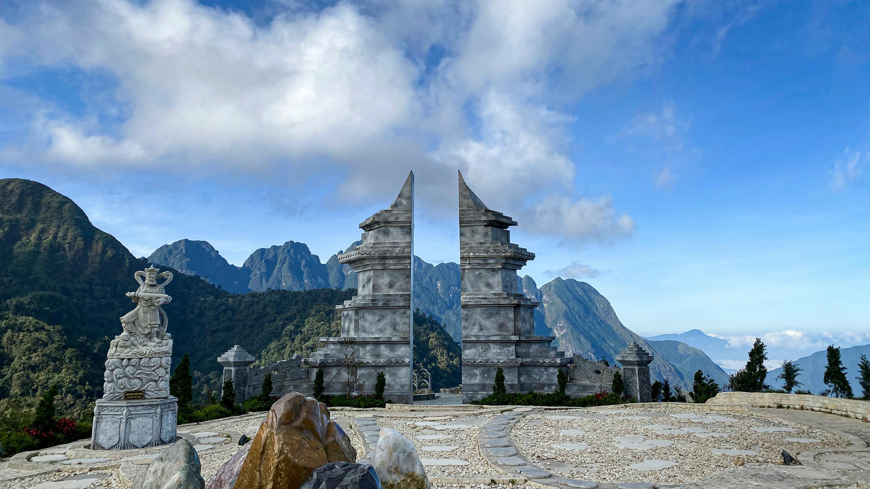 A stone gate with two tall, split pillars on a mountain landscape under a partly cloudy blue sky.