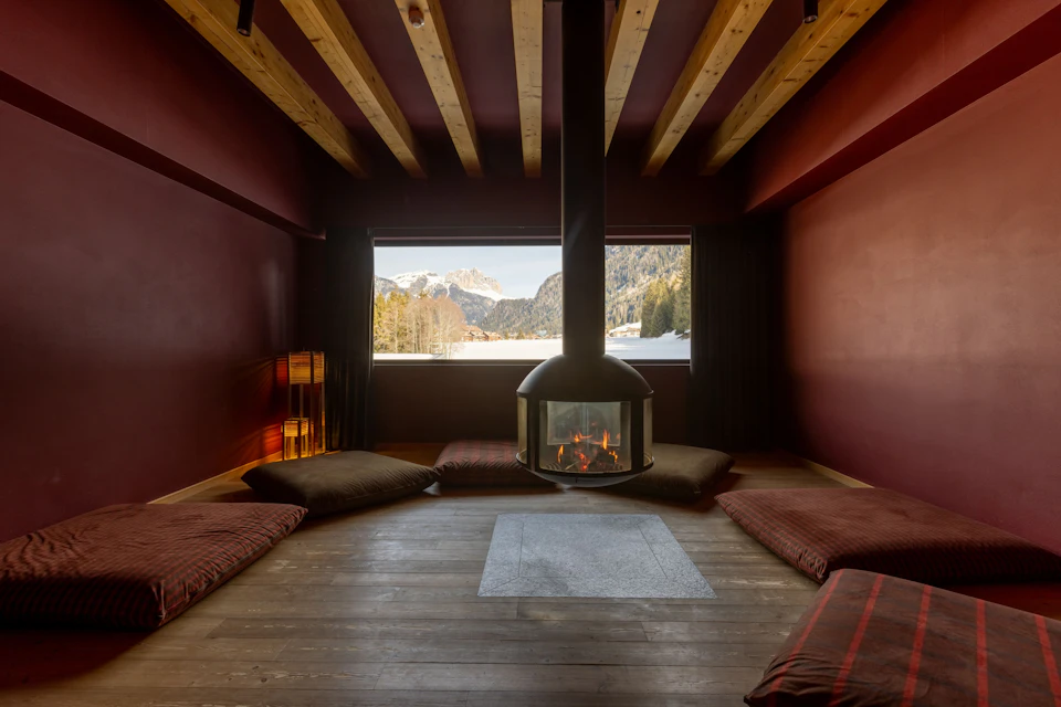 A cozy room with a central suspended fireplace, cushions on the floor, wooden ceiling beams, and a mountain view through a window.