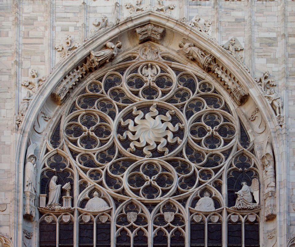 Intricate Gothic-style rose window with ornate stone carvings and statues, set in an arched frame on a stone building facade.