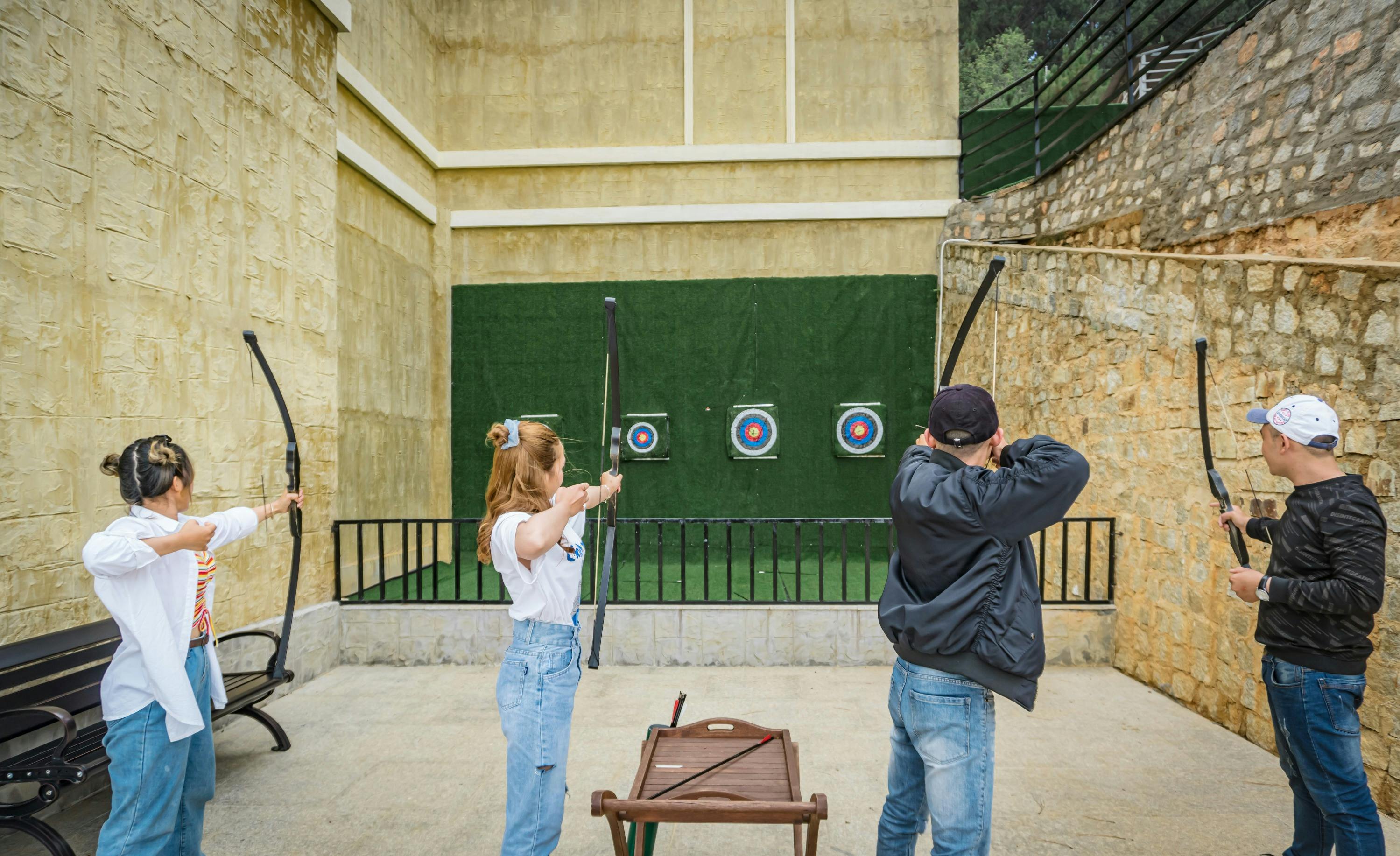 People practicing archery at an outdoor range with three target boards against a green backdrop.