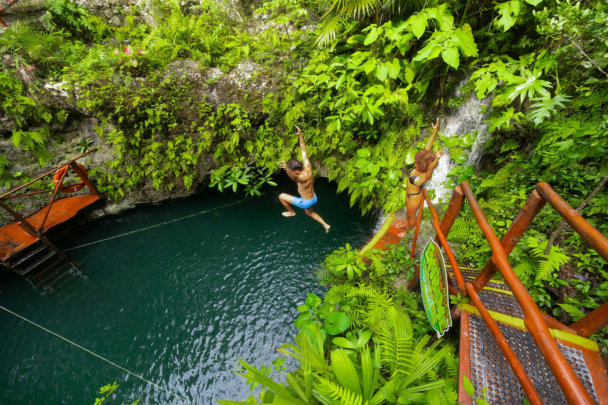 Dues persones salten a un exuberant cenote de la selva des d'una plataforma de fusta, envoltades d'un folre verd vibrant i penya-segats rocosos.