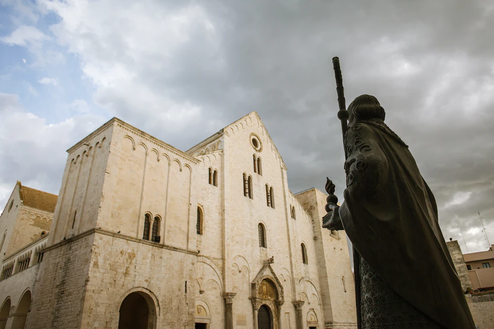 A statue holding a staff stands before a large, historic stone building under a cloudy sky.