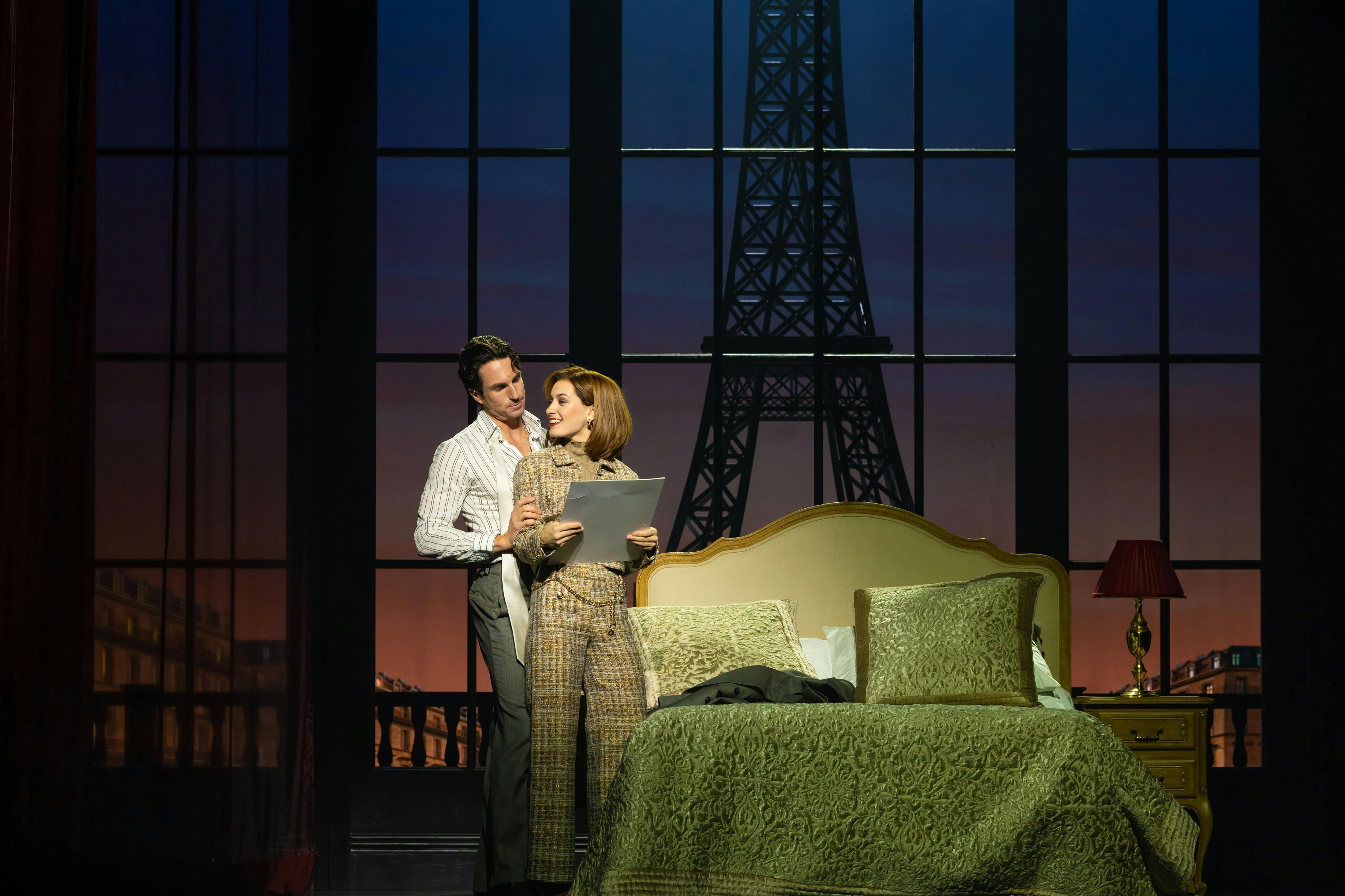 一对夫妇站在一间卧室里，后面有一扇大窗户，展示了埃菲尔铁塔。 一个人拿着文件。