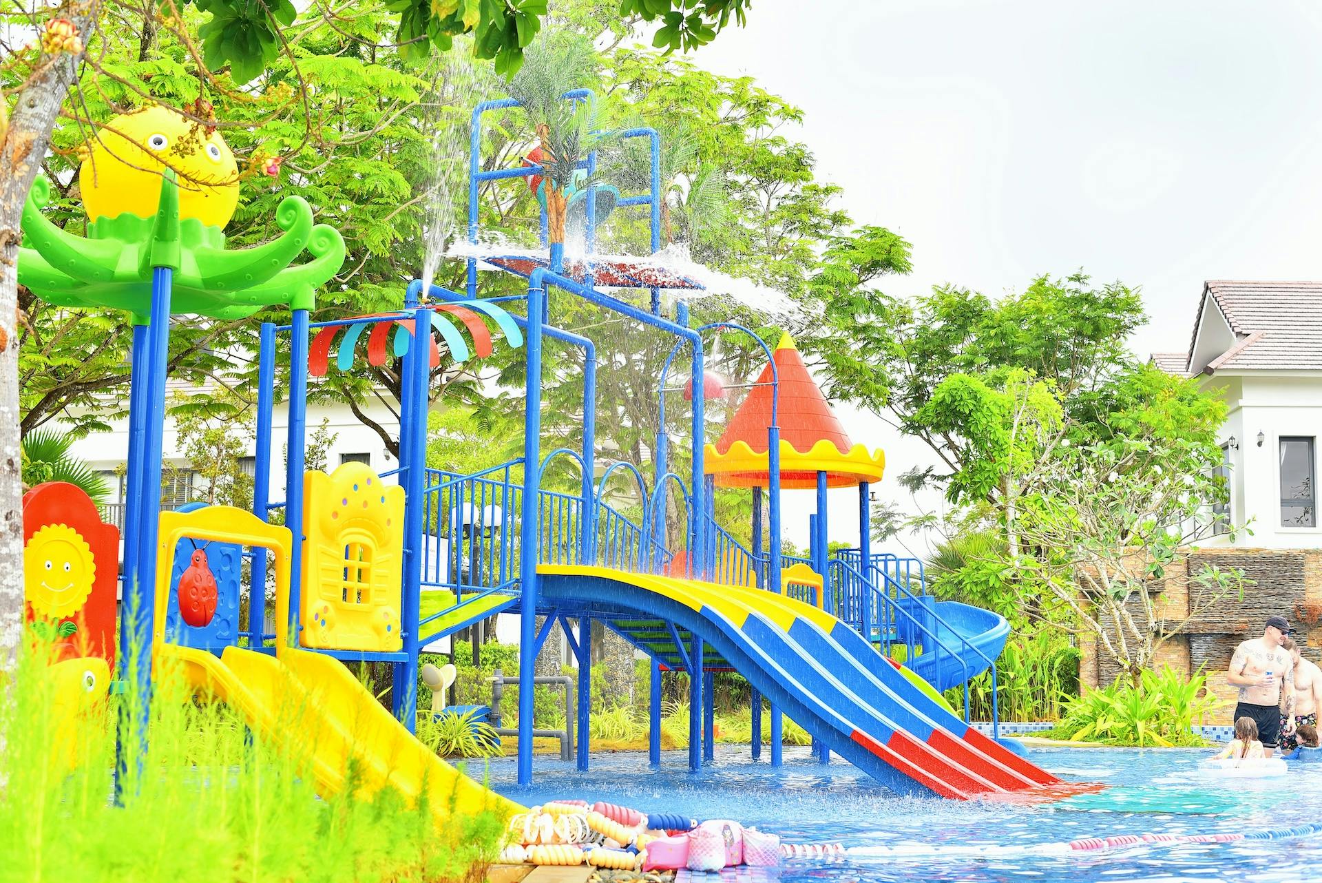 A colorful water playground with slides, climbing structures, and spraying water features, surrounded by trees.
