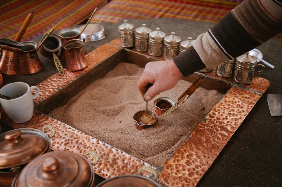 A person preparing coffee using a traditional sand brewing method, with copper pots and containers on a textured tray.