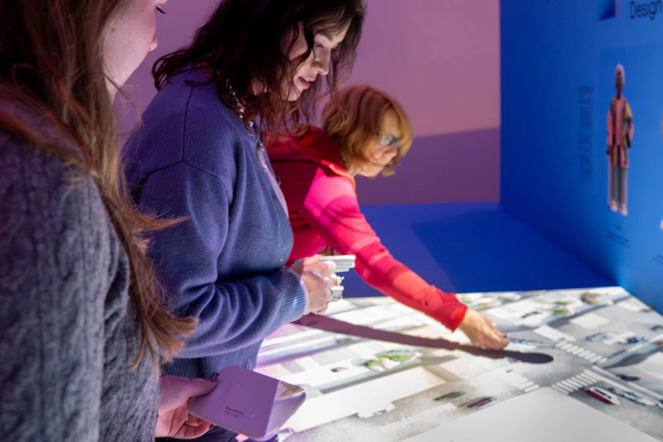 Two women examining an illuminated tabletop display featuring maps and diagrams in a brightly lit room.